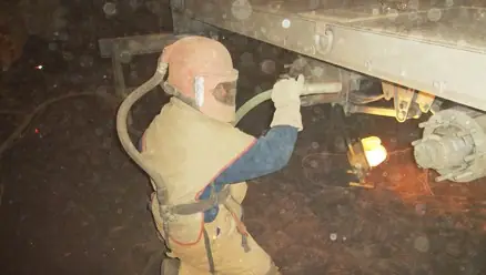 Worker in a blast suit and hood blasting underneath a trailer